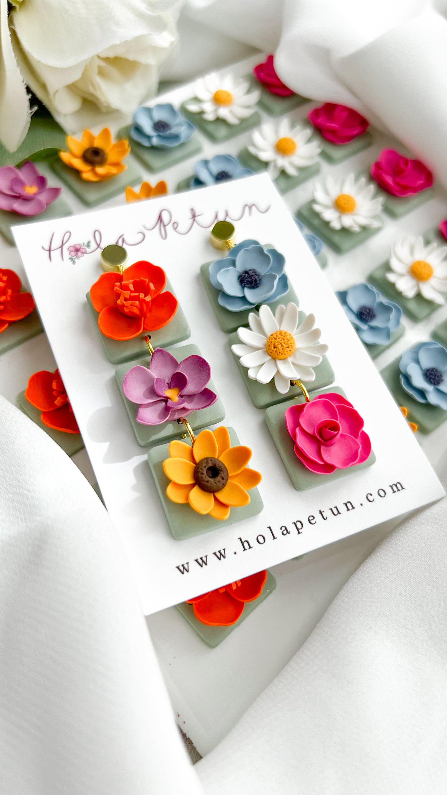 Colorful flower-shaped earrings on a Hola Petun branded card with flowers in the background.