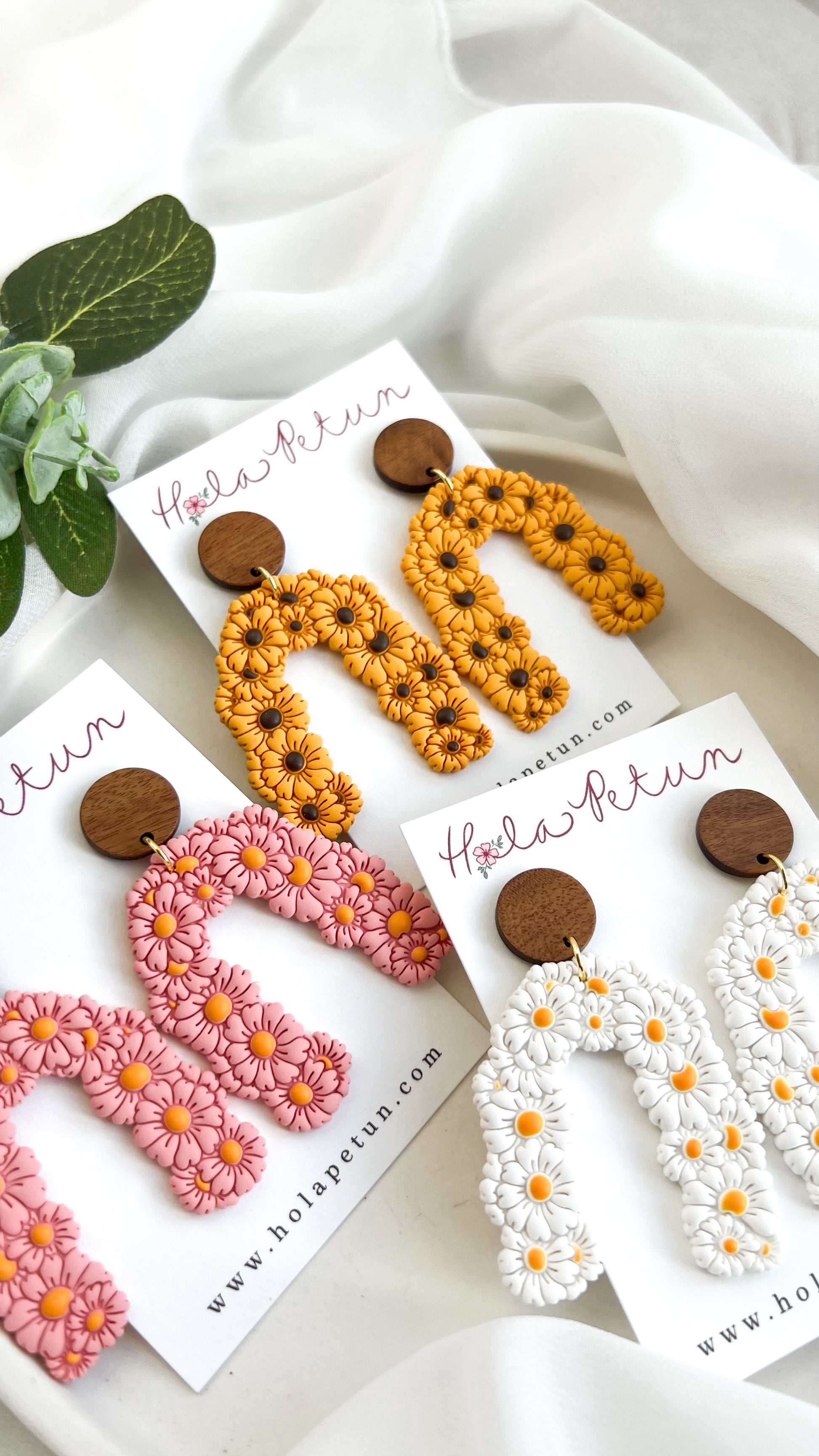 Three pairs of earrings on cards with 'Hola Perin' branding, displayed on a white fabric background.