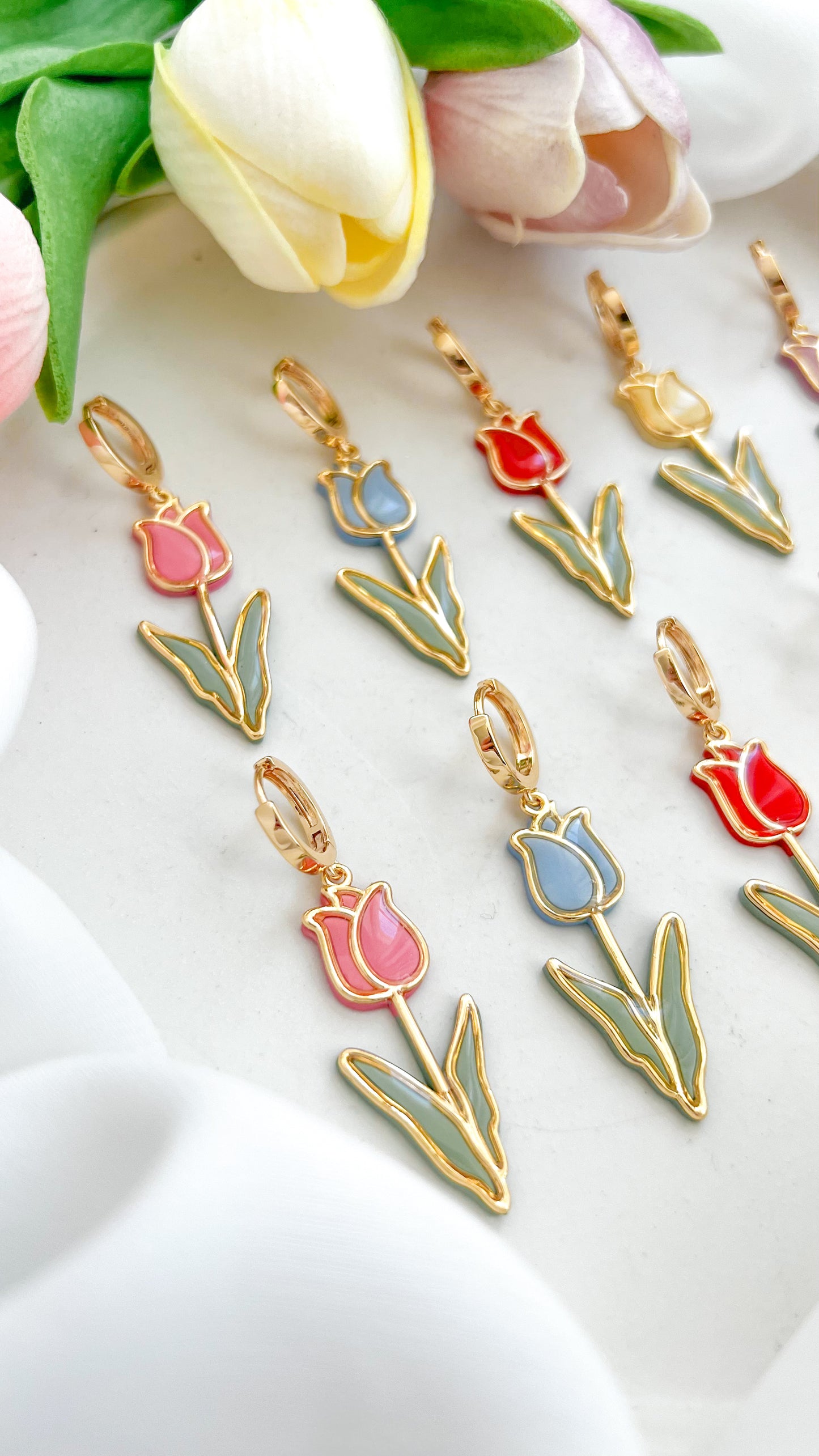 Set of tulip-shaped polymer clay earrings in various colors, pink, blue, red, yellow and purple on a white fabric background with tulips.