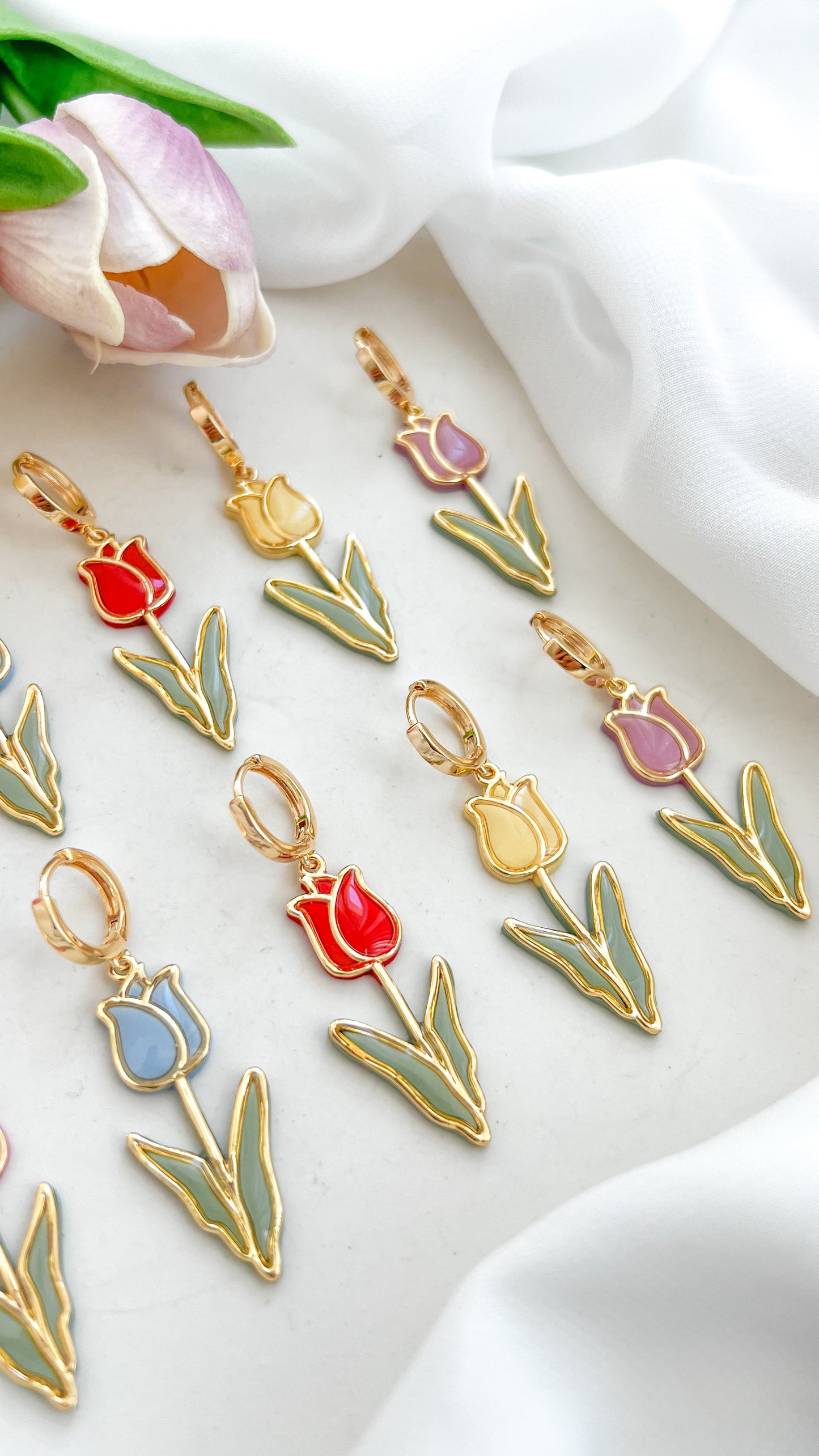 Set of tulip-shaped polymer clay earrings in various colors, pink, blue, red, yellow and purple on a white fabric background with tulips.