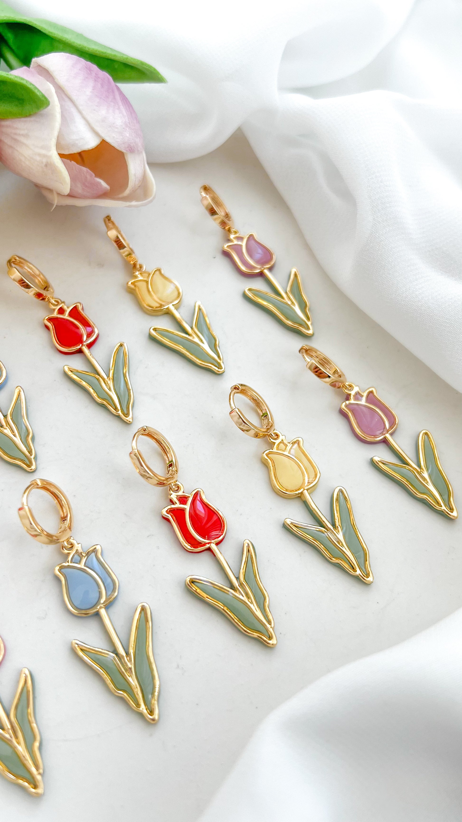 Set of tulip-shaped polymer clay earrings in various colors, pink, blue, red, yellow and purple on a white fabric background with tulips.