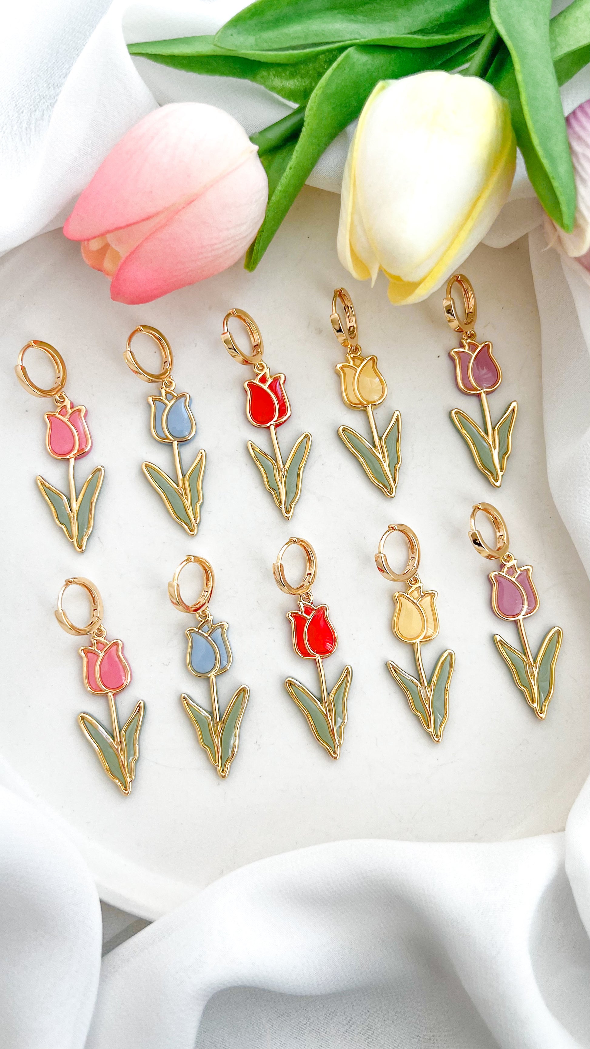 Set of tulip-shaped polymer clay earrings in various colors, pink, blue, red, yellow and purple on a white fabric background with tulips.